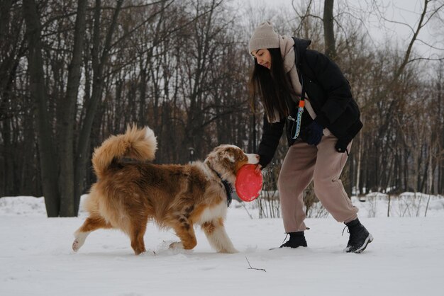 Jonge brunette vrouw loopt in het besneeuwde winterpark met hond en speelt met rode schijf speelgoed