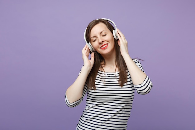 Jonge brunette vrouw meisje in casual gestreepte kleding poseren geïsoleerd op violet paarse achtergrond studio portret. Mensen oprechte emoties lifestyle concept Mock-up kopieerruimte Luister muziek in een koptelefoon