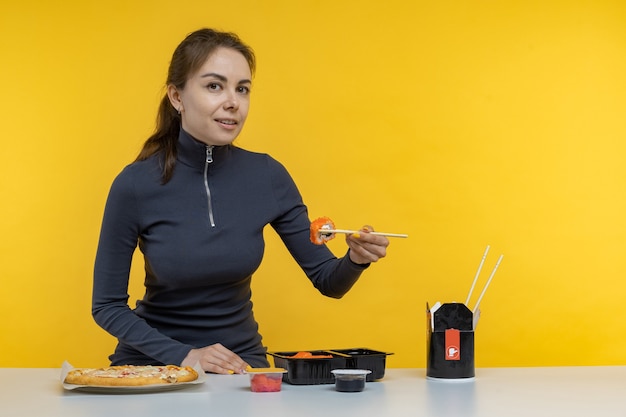 Jonge brunette vrouw meisje in casual kleding houden in de hand makizushi sushi roll geserveerd op zwarte plaat traditionele japanse gerechten geïsoleerd op gele achtergrond studio portret. mensen levensstijl concept