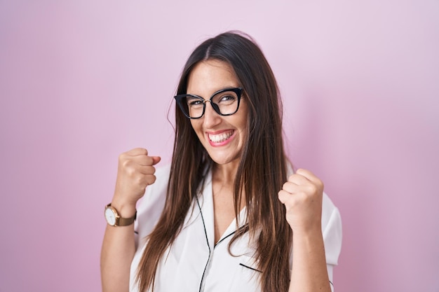 Jonge brunette vrouw met bril staande over roze achtergrond erg blij en opgewonden winnaar gebaar doen met opgeheven armen glimlachend en schreeuwend voor succes viering concept