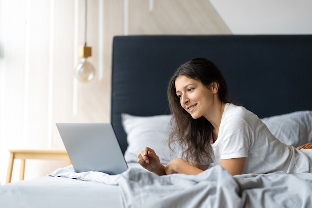 jonge brunette vrouw met een laptop op het bed liggen. Stijlvol modern interieur. Een gezellige werkplek. Winkelen op internet.