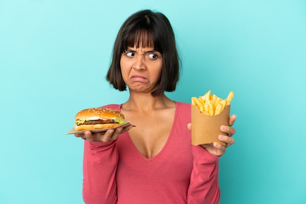 Jonge brunette vrouw met hamburger en gebakken chips over geïsoleerde blauwe achtergrond