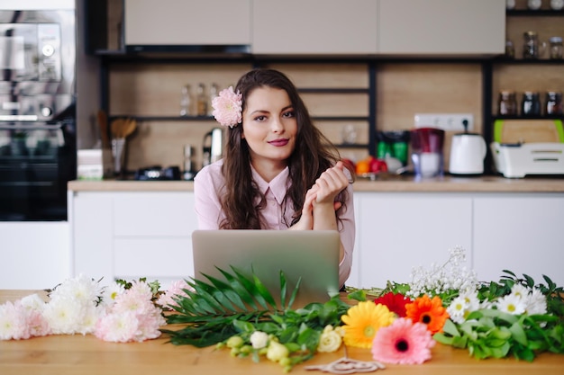 Jonge brunette vrouw met lang golvend haar die op laptop met bloemen op het bureau werkt met keuken op de achtergrond Getalenteerde bloemist die online verkoop ontwikkelt en zich klaarmaakt voor workshop