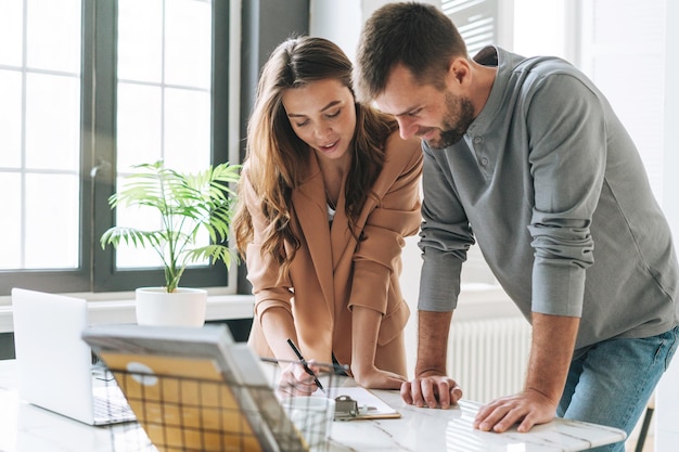 Jonge brunette vrouw met lang haar in stijlvol pak werken met jonge man met behulp van op laptop op tafel met groene kamerplant in het lichte moderne kantoor