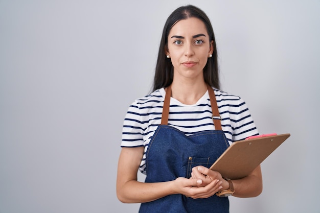 Jonge brunette vrouw met professionele serveersterschort en klembord-denkende houding en nuchtere uitdrukking die er zelfverzekerd uitziet