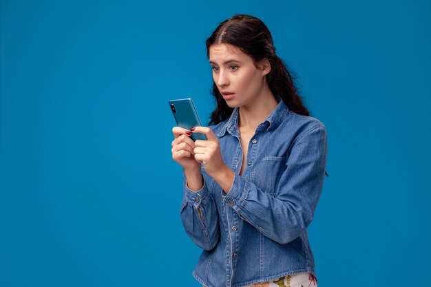 Jonge brunette vrouw poseert met een smartphone die op een blauwe achtergrond staat.