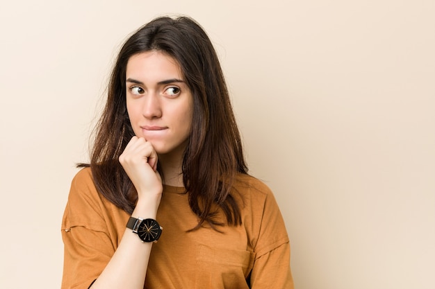 Jonge brunette vrouw tegen een beige achtergrond verward, voelt zich twijfelachtig en onzeker.