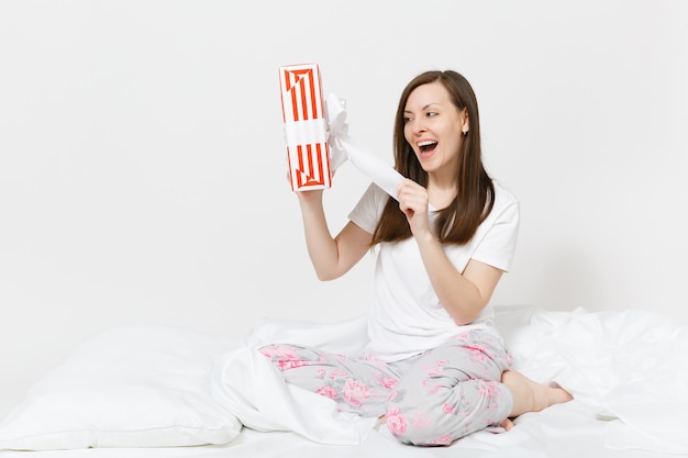 Jonge brunette vrouw zittend in bed met rood gestreepte geschenkdoos, wit laken, kussen, inwikkeling in deken geïsoleerd op een witte muur