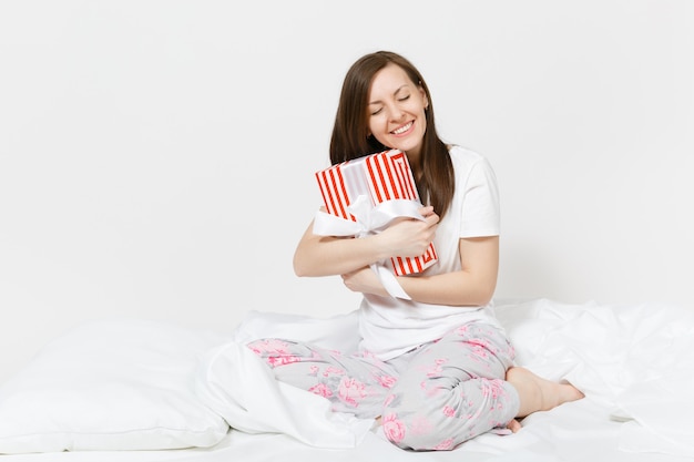 Jonge brunette vrouw zittend in bed met rood gestreepte geschenkdoos, wit laken, kussen, inwikkeling in deken geïsoleerd op een witte muur