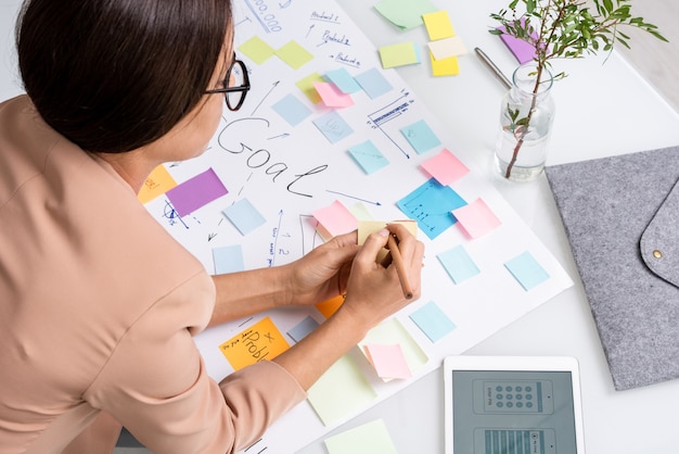 Jonge brunette zakenvrouw die aantekeningen maakt op briefpapier terwijl ze over een groot document buigt met de belangrijkste doelen en werkpunten op de werkplek