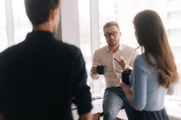 Jonge business team ideeën delen tijdens koffiepauze Twee knappe jonge zakenlieden en dame in casual kleding houden kopjes koffie praten en glimlachen permanent binnenshuis het kantoorgebouw