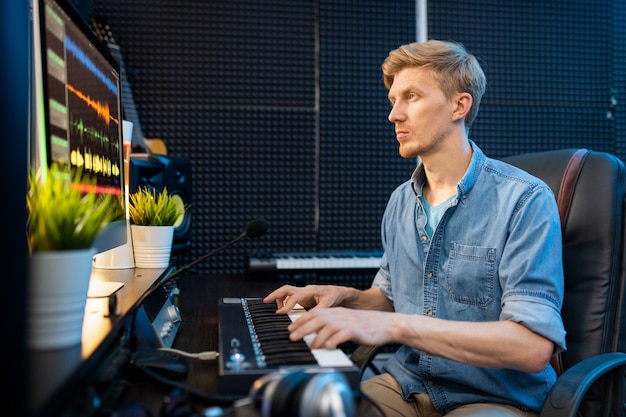 Jonge casual blonde man kijken naar computerscherm tijdens het indrukken van toetsen van piano klavier in geluidsopnamestudio