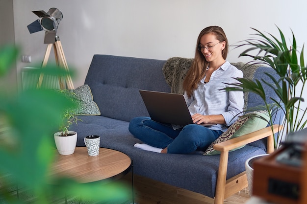 Jonge casual gelukkig lachende vrouw freelancer in ronde glazen zittend op de Bank en online werken op een laptop in comfortabel gezellig huis tussen planten binnenshuis