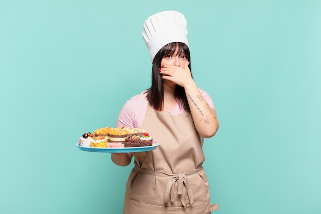 Jonge chef-kokvrouw die mond bedekt met handen met een geschokte, verbaasde uitdrukking, een geheim houdt of oeps zegt