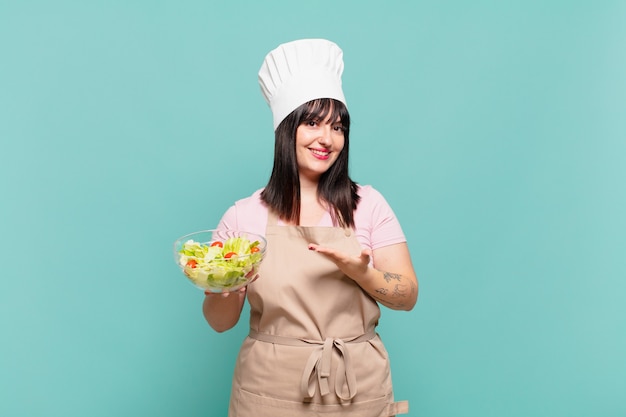 Jonge chef-kokvrouw die vrolijk lacht, zich gelukkig voelt en een concept toont in kopieerruimte met handpalm