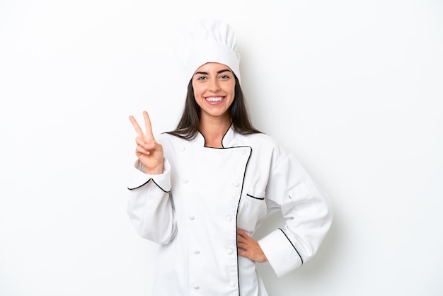 Jonge chef-kokvrouw over witte achtergrond die en overwinningsteken glimlacht toont