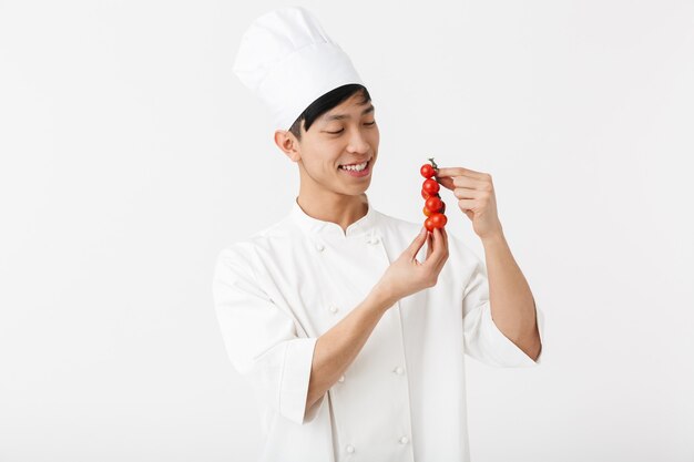 jonge chinese chef-man in het wit kok uniform glimlachend in de camera terwijl tomatengroenten geïsoleerd over witte muur