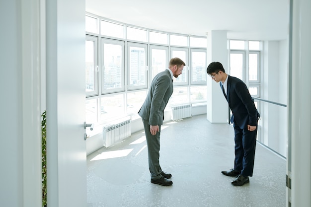 Jonge Chinese en Kaukasische zakenlieden in formalwear die tijdens begroeting naar elkaar buigen terwijl ze in een groot kantoorgebouw staan