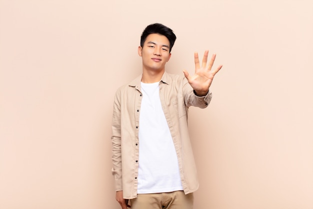 Jonge Chinese en mens die vriendelijk glimlachen kijken, tonend nummer vijf of vijfde met voorwaartse hand, aftellend tegen vlakke kleurenmuur