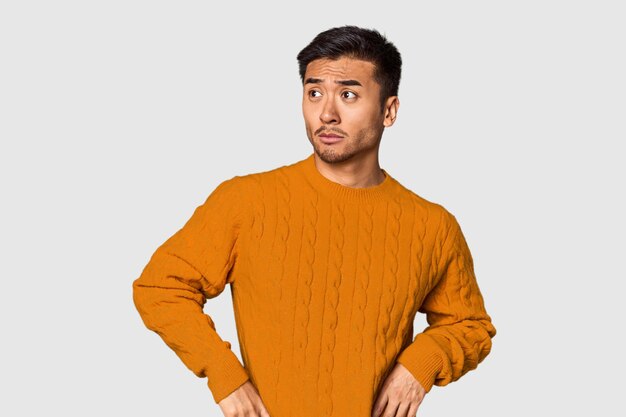 Jonge Chinese man in de achtergrond van de studio verward voelt zich twijfelachtig en onzeker