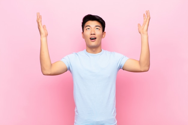 Jonge Chinese mens die gelukkig, verbaasd, gelukkig en verrast voelt, die overwinning met beide handen omhoog in de lucht tegen vlakke kleurenmuur viert