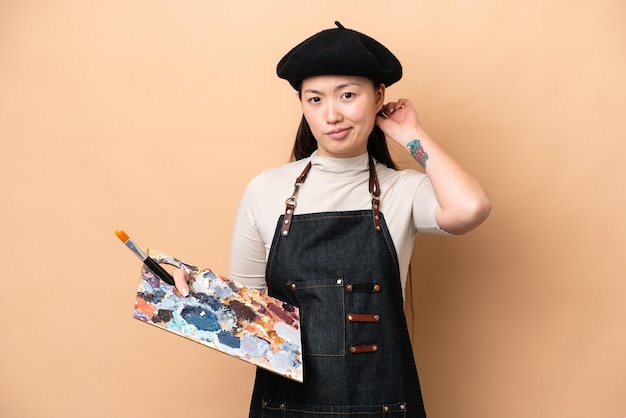 Jonge Chinese schildersvrouw die op beige achtergrond wordt geïsoleerd die twijfels heeft