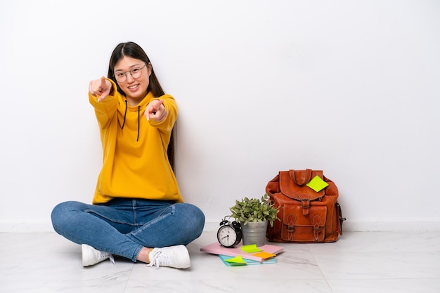 Jonge Chinese studente vrouw zittend op de vloer geïsoleerd op een witte muur wijst vinger naar je terwijl ze lacht