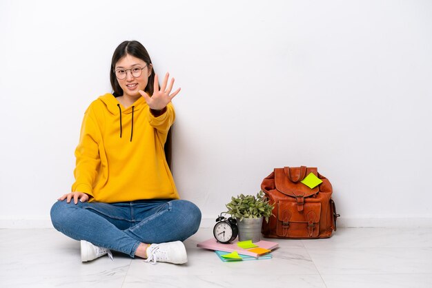 Jonge Chinese studentenvrouw zittend op de vloer geïsoleerd op een witte muur die vijf telt met vingers