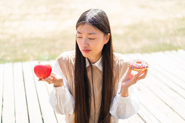 Jonge Chinese vrouw die buitenshuis appel en donut vasthoudt en nadenkt