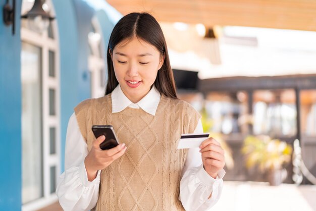 Jonge Chinese vrouw die buitenshuis koopt met de mobiel met een creditcard