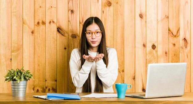 Jonge Chinese vrouw die op haar bureau bestudeert dat lippen vouwt en palmen houdt om luchtkus te verzenden.