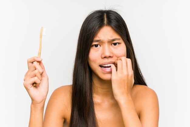 Jonge Chinese vrouw die tandenborstel geïsoleerde het bijten vingernagels, zenuwachtig en zeer bezorgd houdt.