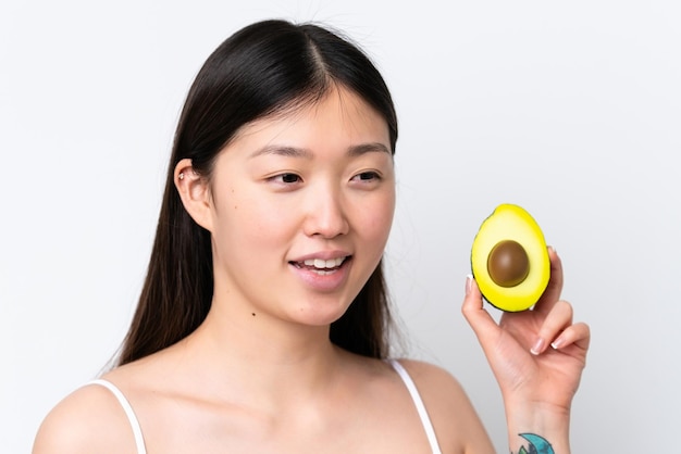 Jonge Chinese vrouw geïsoleerd op een witte achtergrond die een avocado vasthoudt terwijl ze lacht Close-up portret