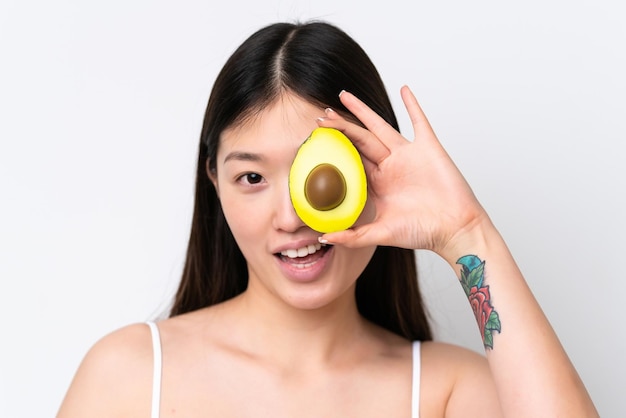 Jonge Chinese vrouw geïsoleerd op een witte achtergrond die een avocado vasthoudt terwijl ze lacht Close-up portret