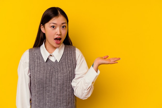 Jonge chinese vrouw geïsoleerd op gele achtergrond onder de indruk de ruimte van het bedrijfskopie op palm.