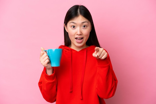 Jonge Chinese vrouw met kopje koffie geïsoleerd op roze achtergrond verrast en wijzend naar voren