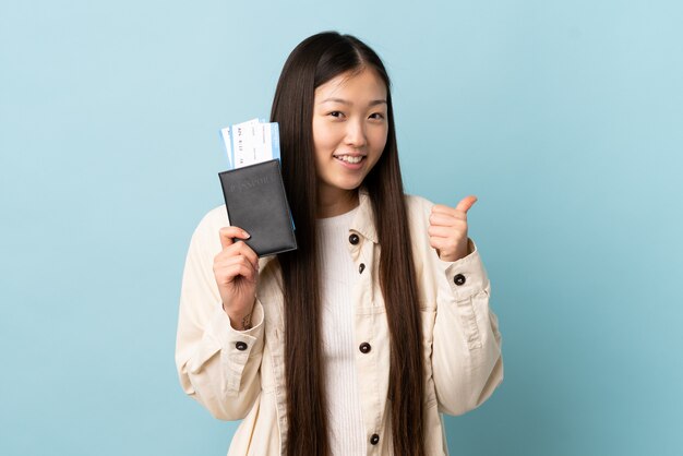 Jonge Chinese vrouw over geïsoleerde muur in vakantie die een paspoort en een vliegtuig met omhoog duim houdt