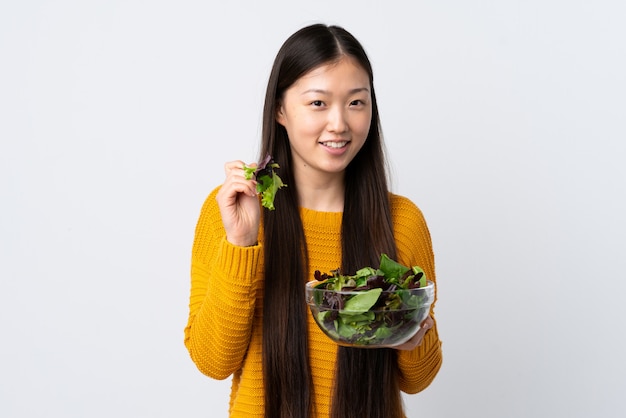 Jonge Chinese vrouw over geïsoleerde witte muur die een kom salade met gelukkige uitdrukking houdt