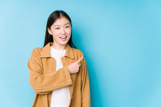 Jonge Chinese vrouw poseren in een blauwe muur geïsoleerd glimlachend en opzij wijzend, iets laten zien op lege ruimte.