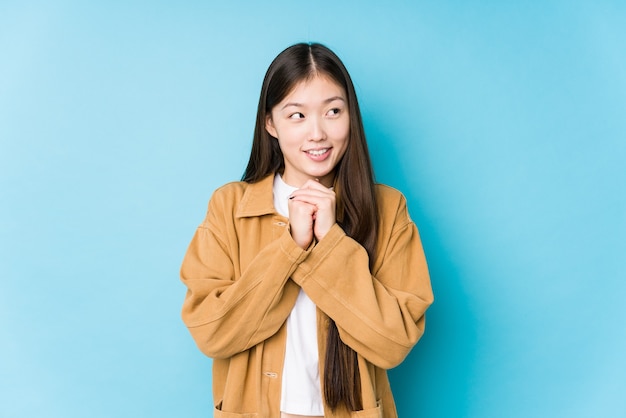 Jonge Chinese vrouw poseren in een blauwe muur geïsoleerd houdt handen onder de kin, kijkt gelukkig opzij.