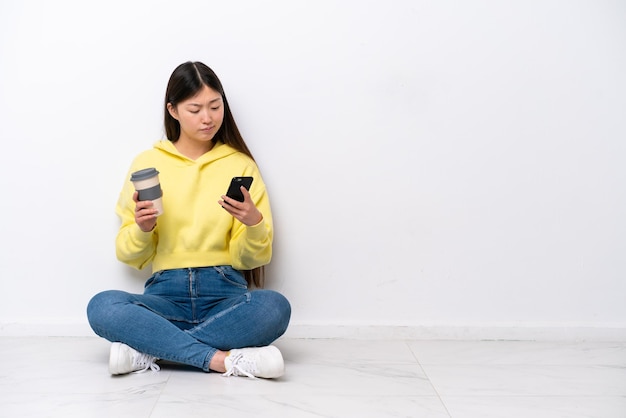 Jonge Chinese vrouw zittend op de vloer geïsoleerd op een witte muur met koffie om mee te nemen en een mobiel