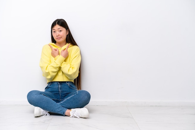 Jonge Chinese vrouw zittend op de vloer geïsoleerd op een witte muur wijzend naar de zijkanten met twijfels