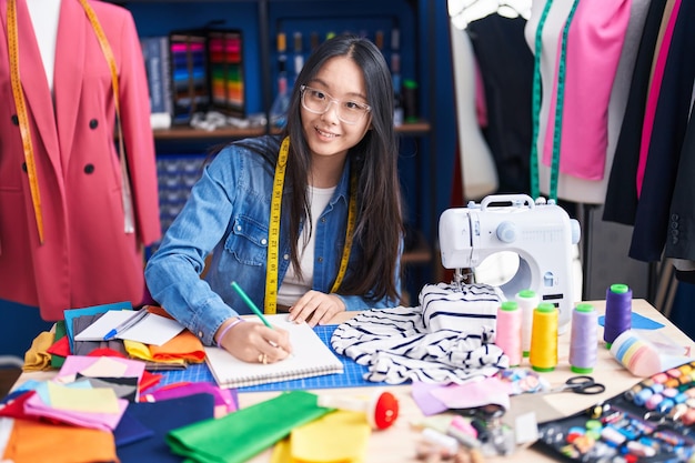 Jonge chinese vrouwenkleermaker glimlachend zelfverzekerd tekenend op een notitieboekje in de kledingfabriek