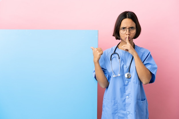 Jonge chirurgenvrouw van gemengd ras met een groot spandoek over een geïsoleerde achtergrond die naar de zijkant wijst en een stiltegebaar doet