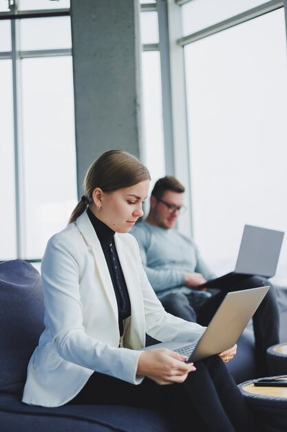 Jonge collega's zitten op zachte poefs en werken op afstand op laptops Moderne werkruimte met grote ramen