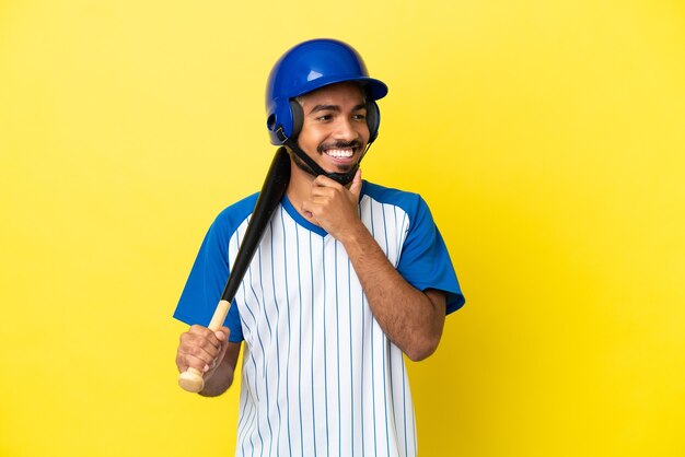 Jonge Colombiaanse Latijns-man die honkbal speelt geïsoleerd op een gele achtergrond, naar de zijkant kijkt en glimlacht