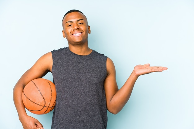 Jonge Colombiaanse man die geïsoleerd basketbal speelt met een kopie ruimte op een handpalm en een andere hand op de taille houdt.