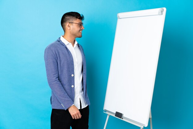 Jonge Colombiaanse man geïsoleerd op blauw met een presentatie op wit bord en op zoek naar kant