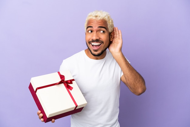 Jonge Colombiaanse man met een geschenk geïsoleerd op een paarse achtergrond, luisterend naar iets door hand op het oor te leggen