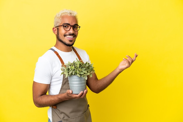 Jonge Colombiaanse man met een plant geïsoleerd op een gele achtergrond die zijn handen naar de zijkant uitstrekt om uit te nodigen om te komen